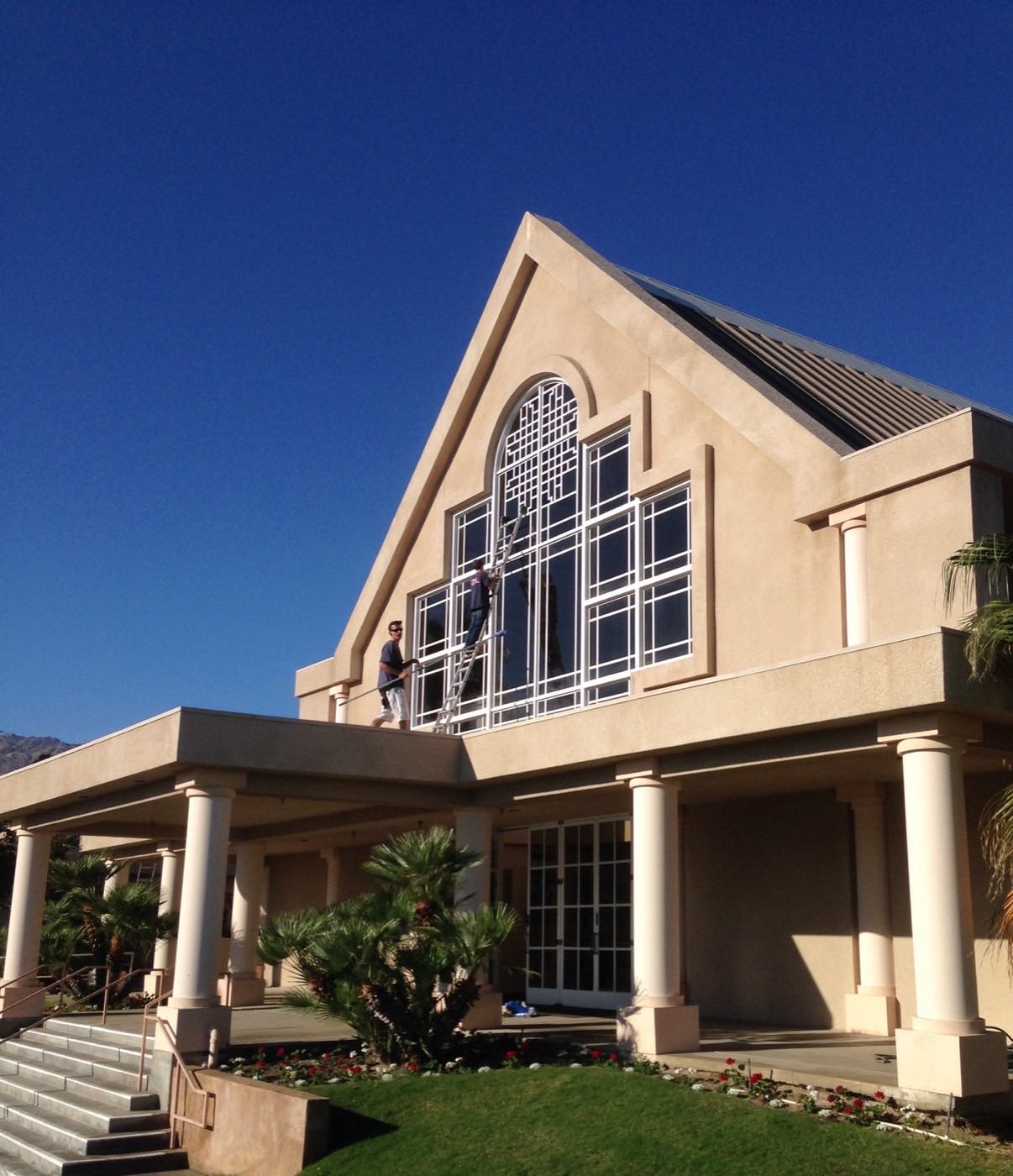 Window cleaning crew on ladders detailing a dramatic two-story arched window wall under a blue desert sky