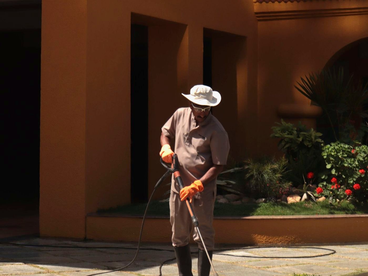 Pressure washing a paved walkway beside a stucco wall and desert planting in strong afternoon sun
